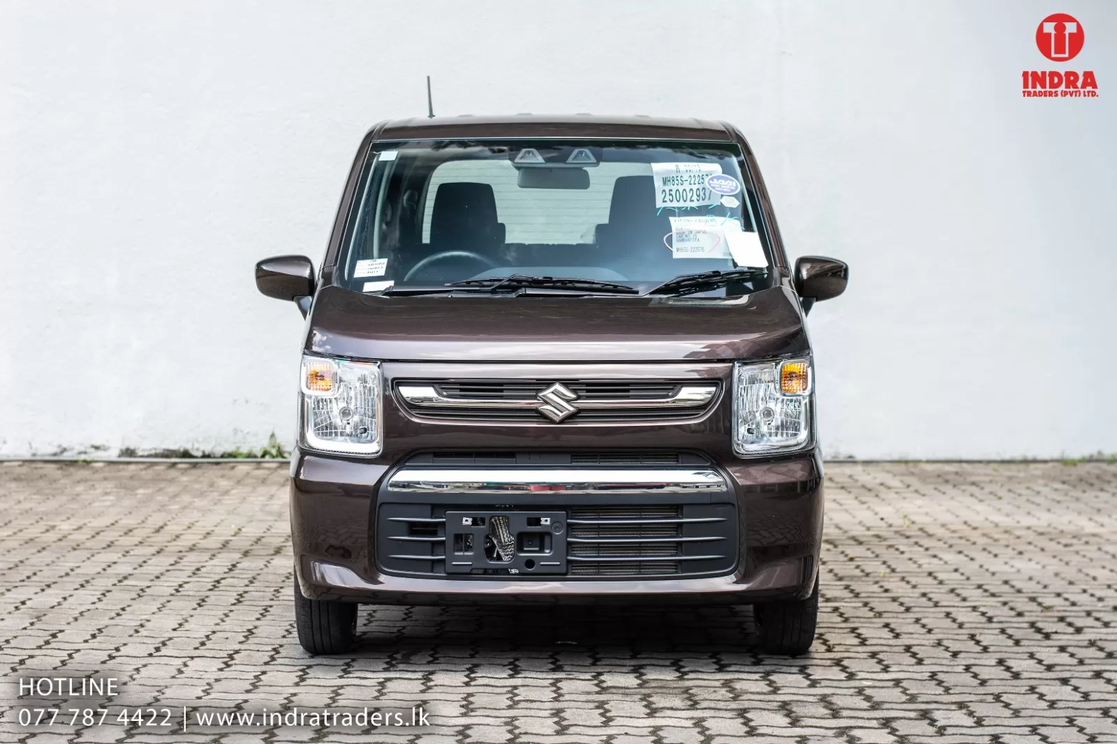 Front view of a brown Suzuki car parked on a brick lot, with a white wall behind and dealership logo at top-right (Indra Traders).