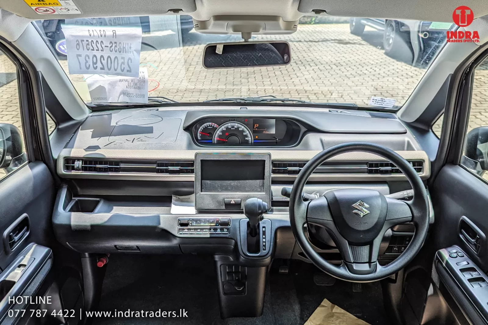 Right-hand drive car interior showing steering wheel with Suzuki logo, central touchscreen, analog gauges, and gear selector.