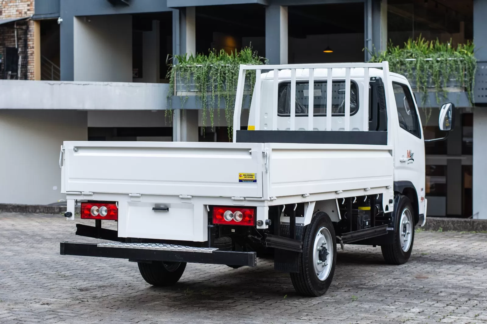 White flatbed utility truck parked on a brick-paved lot, showing the rear cargo bed and red taillights.