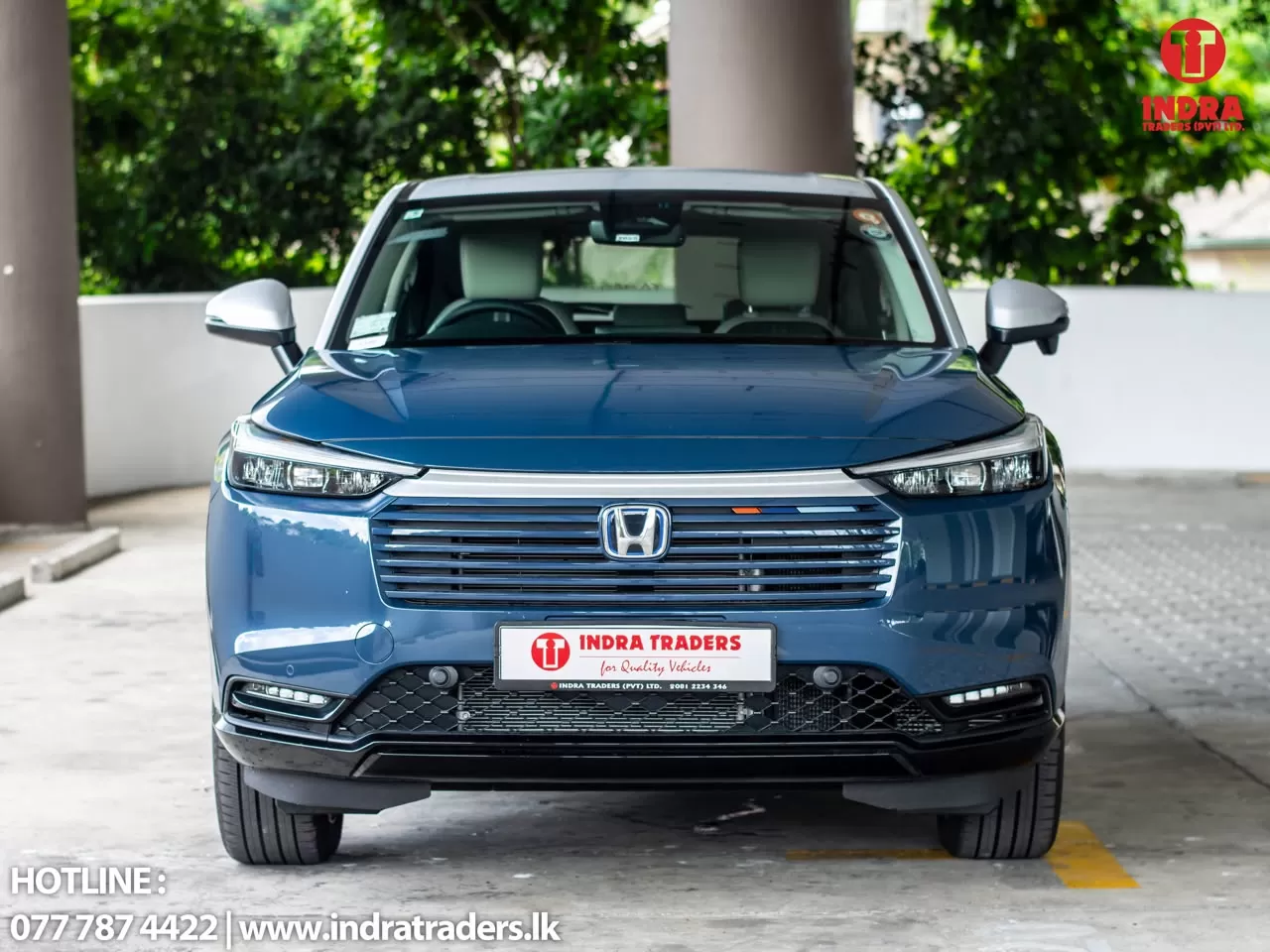Front view of a blue Honda SUV in a parking area, with the Indra Traders branding on the license plate and logo in the top-right corner of the image, and hotline text at the bottom-left.