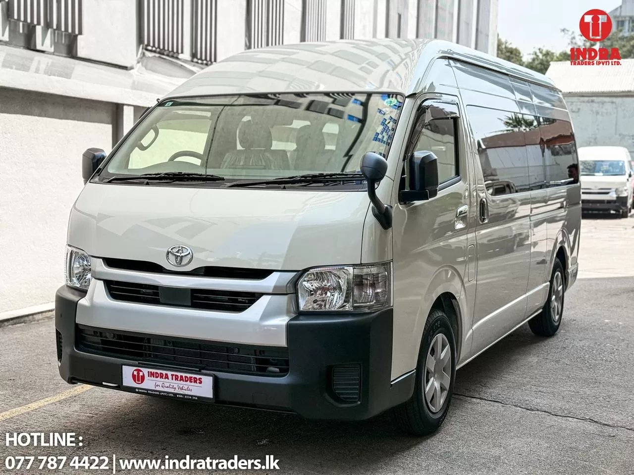 White Toyota van parked in an outdoor lot, front-left angle showing its grille and headlights, with Indra Traders branding visible on the top-right.