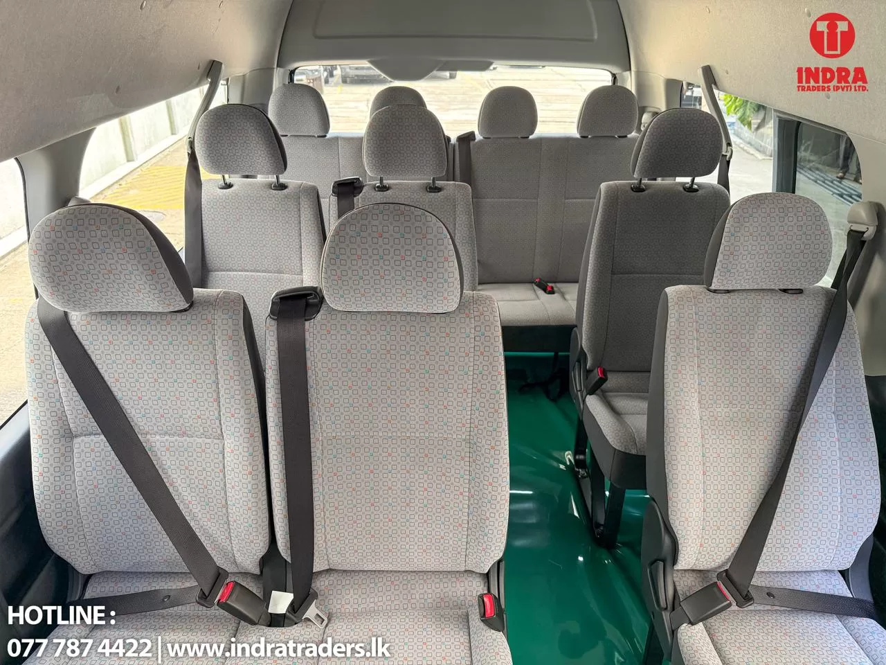 Interior of a gray passenger van showing multiple rows of patterned seats with seat belts and a green floor, Indra Traders logo in top-right corner.