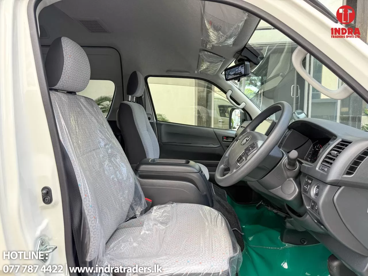 Front interior of a white van showing gray fabric seats with plastic covers, dashboard, and steering wheel; Indra Traders watermark in corner.