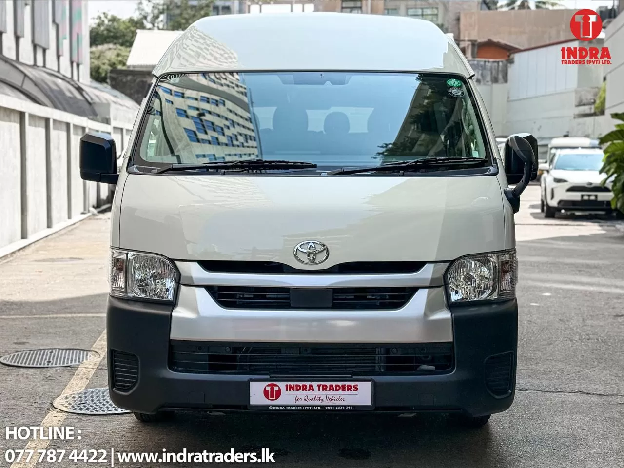Front view of a white Toyota van parked on a city street, with Indra Traders branding on the bumper plate and logo in the top-right corner.