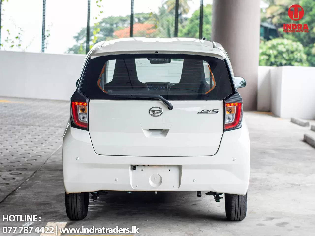 White hatchback rear view parked in a lot, Indra Traders logo in the top-right corner, with hotline and website text at the bottom left.