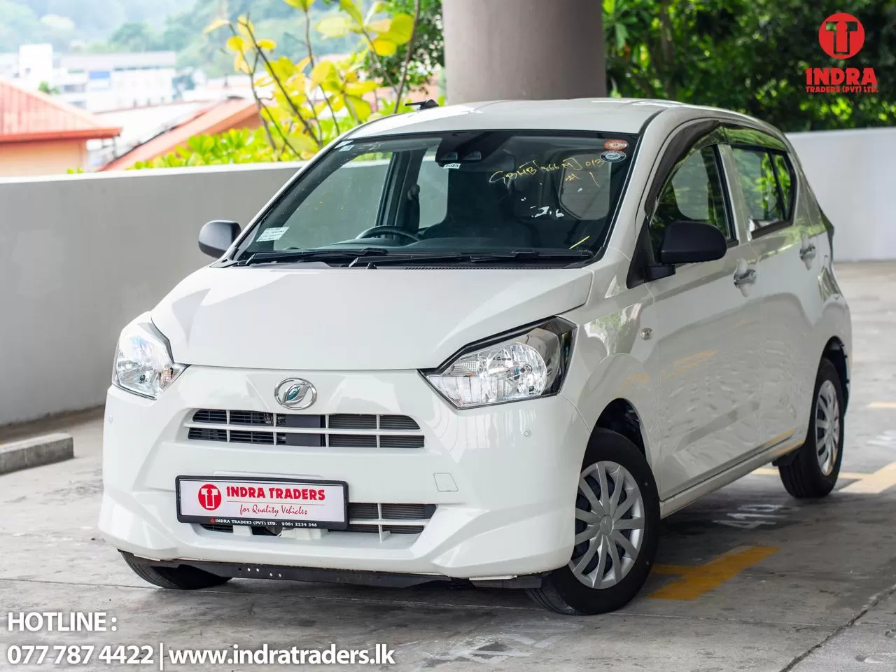 Front three-quarter view of a white compact car parked in a lot, displaying Indra Traders branding on the license plate and a logo in the top-right corner.